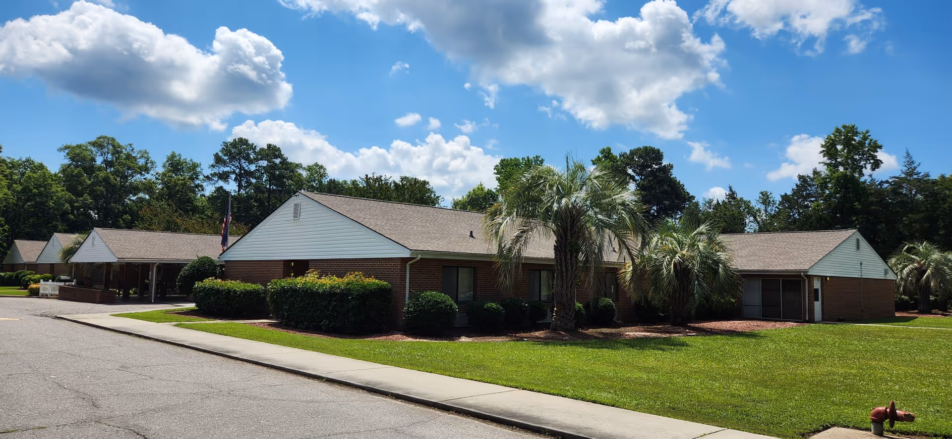 Single-story brick rehabilitation building with palm trees, a manicured lawn and a driveway under a blue sky.
