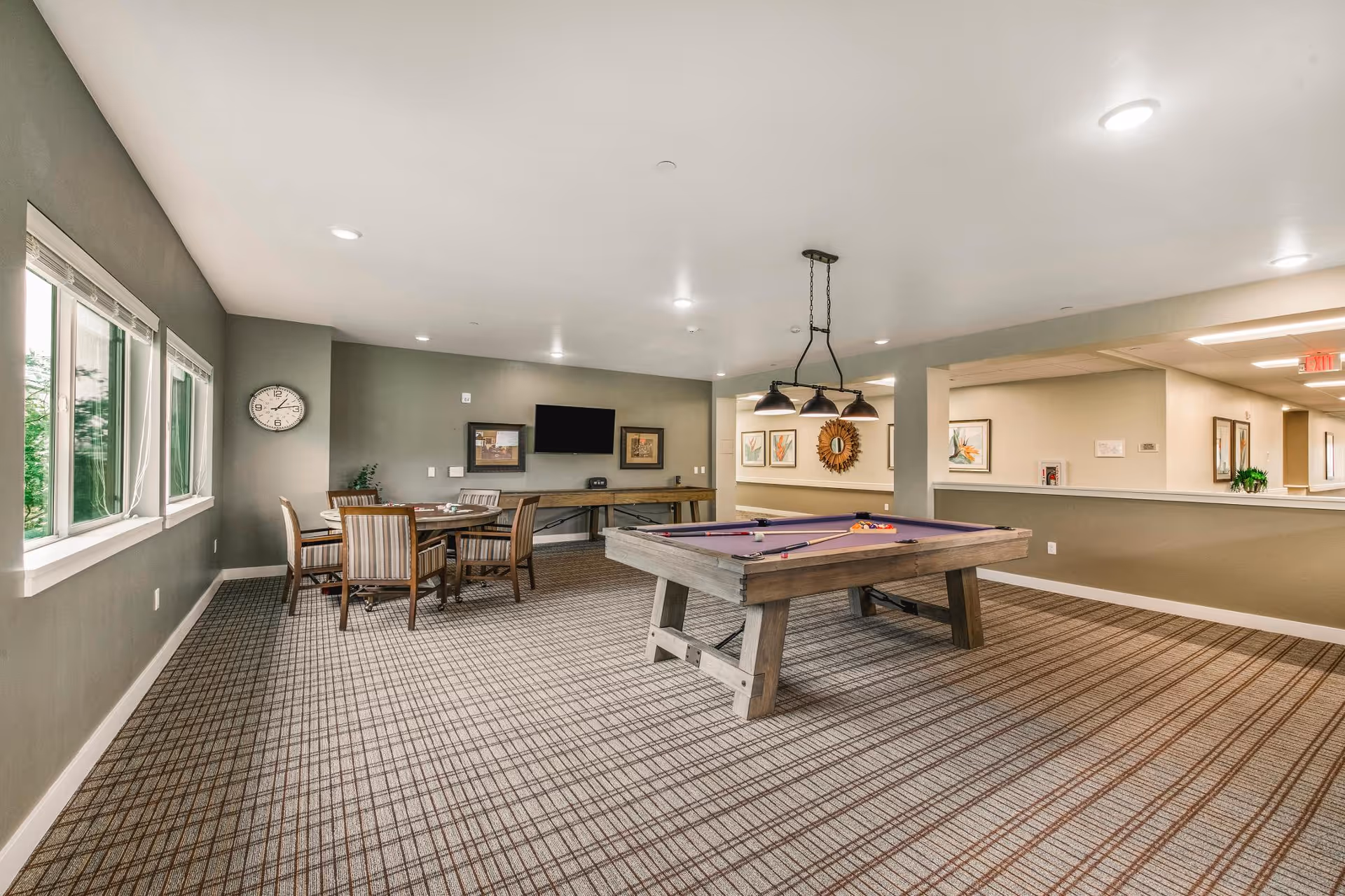 A spacious recreational room with a pool table in the center and a round table with chairs near the windows. The room has a patterned carpet, gray walls, and several framed artworks. There is a wall-mounted TV and a clock on the wall. Ceiling lights and a hanging light fixture illuminate the room.