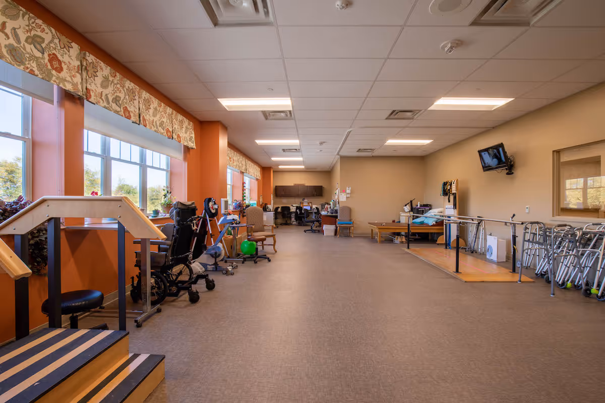 A spacious rehabilitation therapy room with large windows on the left side letting in natural light. The room contains various physical therapy equipment including parallel bars, walkers, wheelchairs, exercise balls, and therapy tables. There are chairs and desks towards the back of the room, and a wall-mounted TV on the right side. The walls are painted in warm tones with floral valances above the windows.