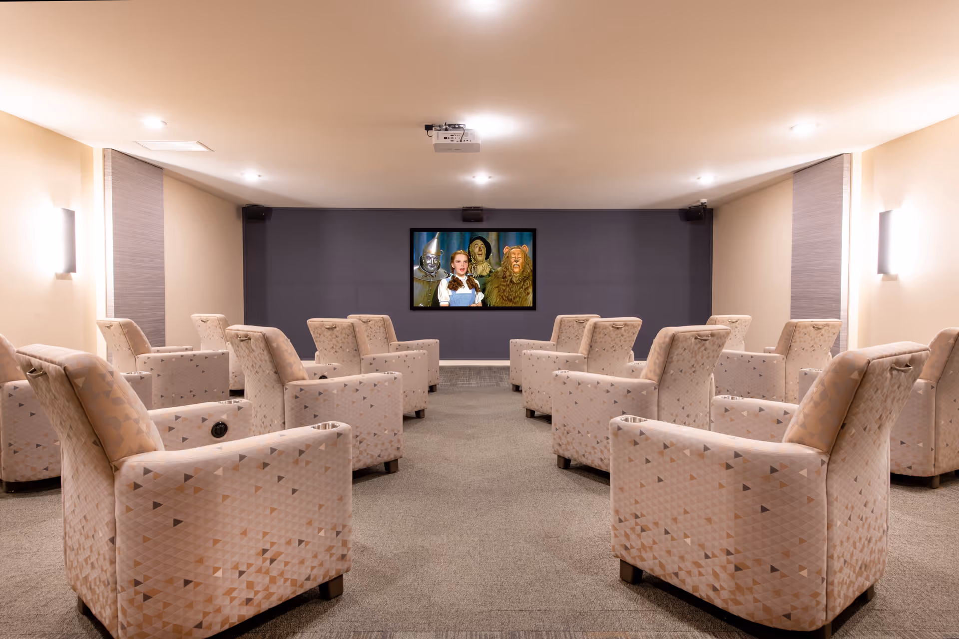 A small screening room with rows of patterned upholstered armchairs facing a wall-mounted TV showing The Wizard of Oz.