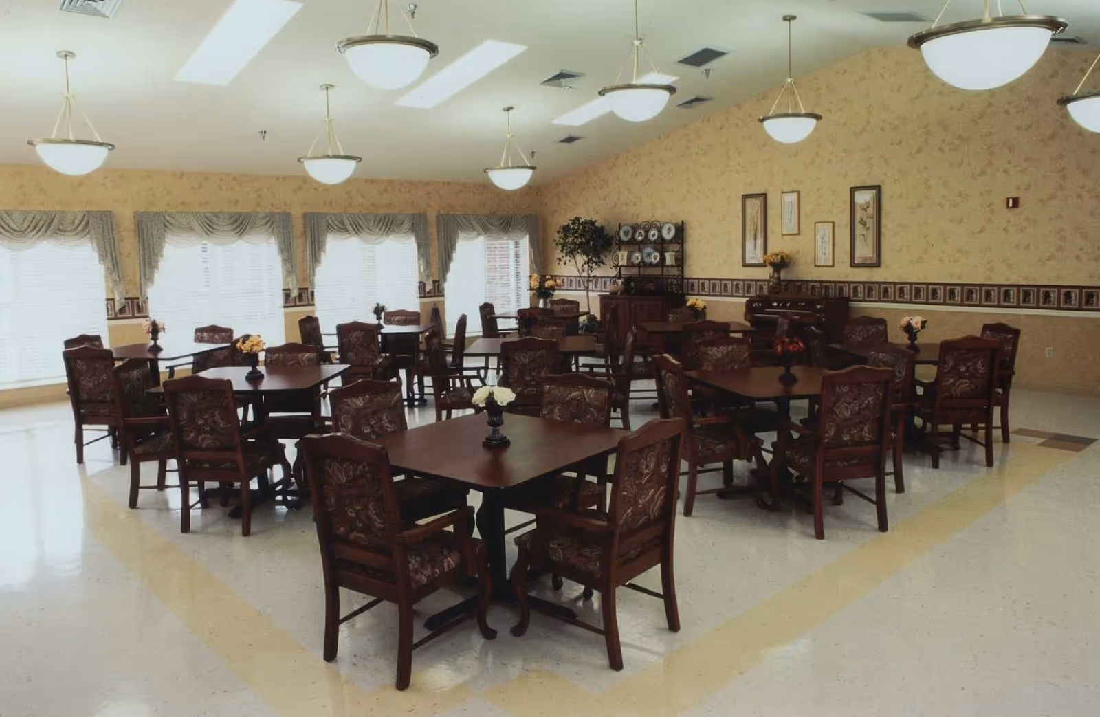 A spacious dining room with multiple dark wood tables and chairs arranged neatly. Each table has a small floral centerpiece. The room has large windows with sheer curtains and valances, floral wallpaper, and several ceiling lights providing bright illumination. Decorative plates and framed artwork adorn the far wall.