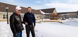 Two men standing outside in a snowy area near a building with beige siding and a brown roof. One man is wearing a black leather jacket and white cap, while the other is wearing a dark jacket and glasses. There is a wooden gazebo and some outdoor furniture visible in the background.
