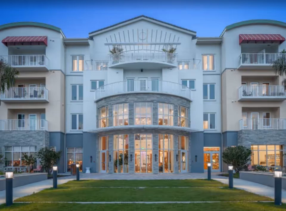 Exterior view of a modern multi-story senior living facility building with balconies, large windows, and a well-maintained lawn with pathway lights in front during dusk.