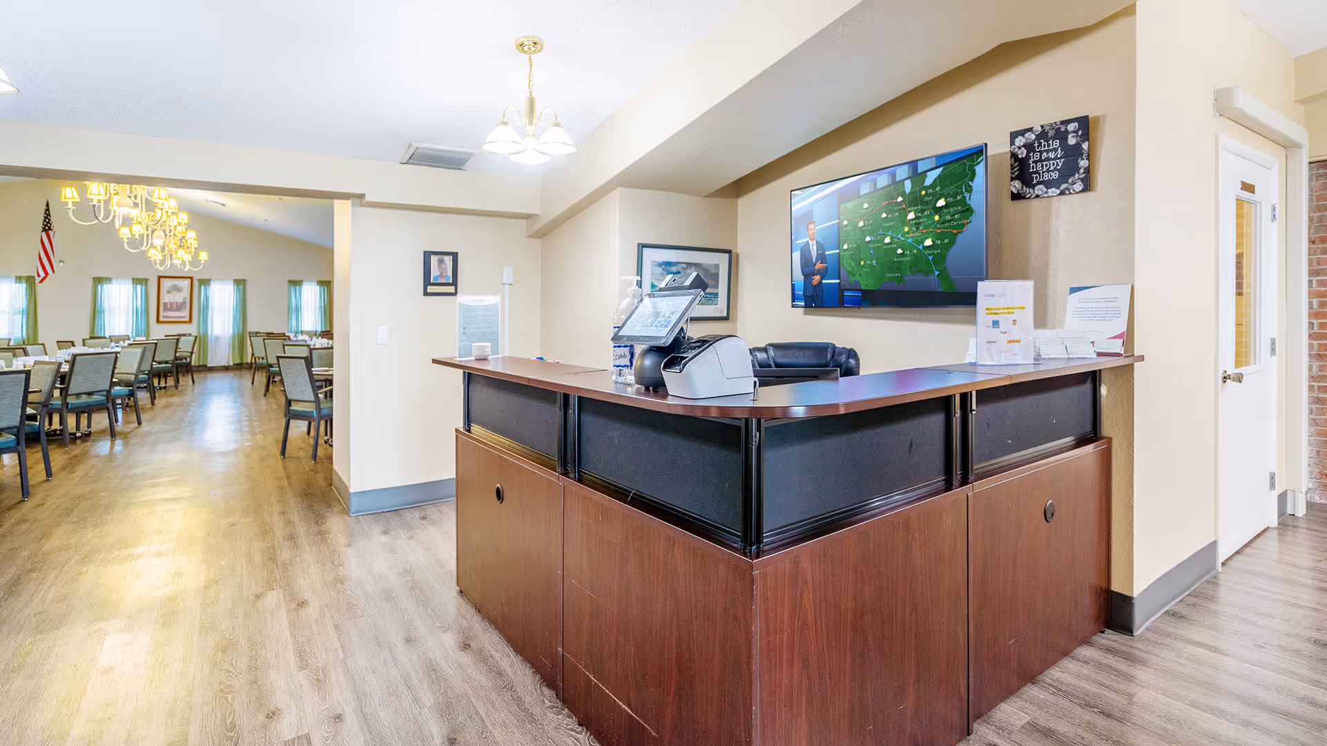 Reception desk area with a computer and chair, adjacent to a dining room with multiple tables and chairs. The dining room has chandeliers, green curtains, and an American flag on the wall. A large TV screen displaying a weather forecast is mounted on the wall behind the reception desk.