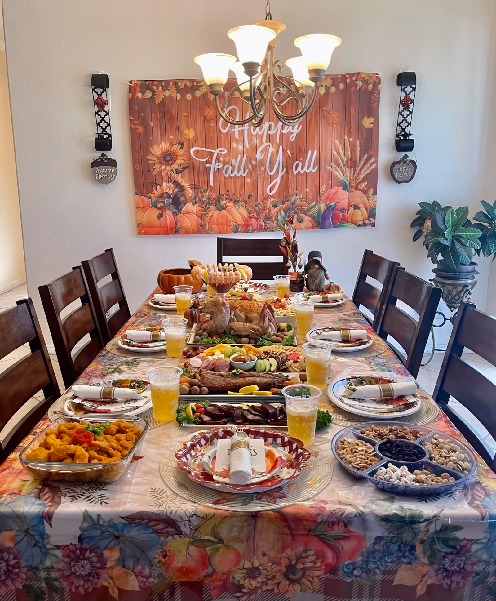 A dining room table set for a festive fall meal with various dishes, drinks, and napkins arranged neatly. The table is covered with a floral and autumn-themed tablecloth. On the wall behind the table is a decorative banner that reads 'Happy Fall Y'all' with pumpkins and autumn leaves. There are wooden chairs around the table and a chandelier overhead. A potted plant is visible to the right side of the image.