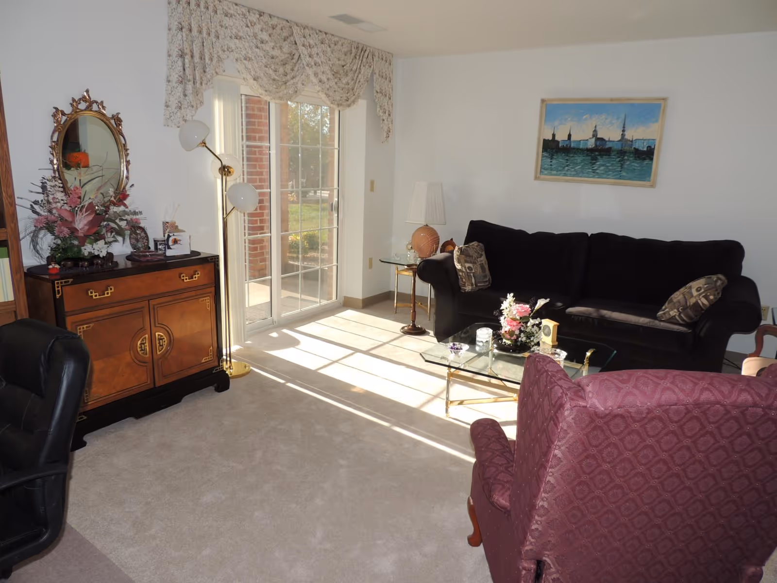 Sunlit living room with a sliding glass door, black sofa, glass coffee table, patterned armchair, and decorative wooden cabinet.