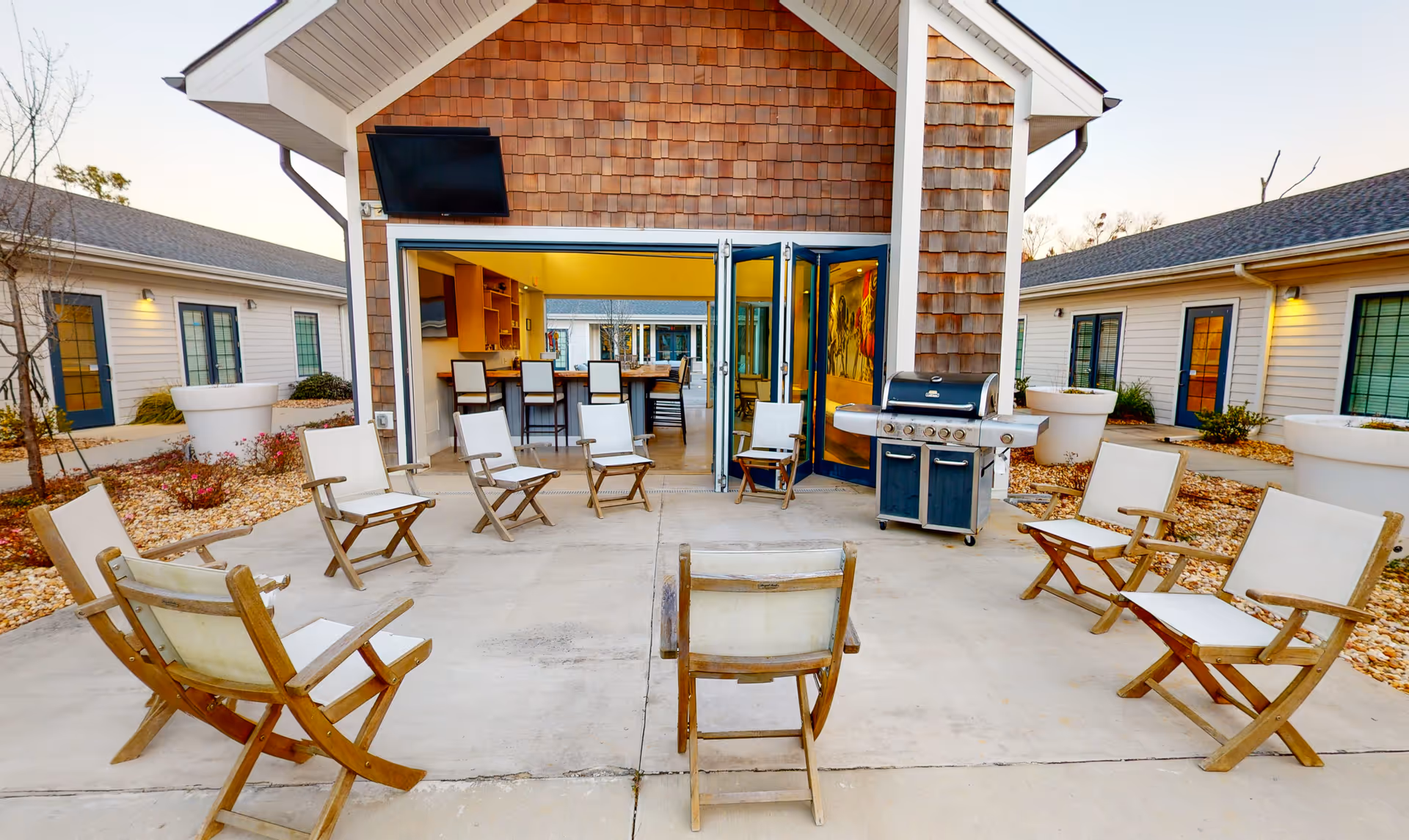 Outdoor patio area with wooden chairs arranged in a circle on a concrete surface. In the background, there is a building with an open folding door revealing a kitchen and dining area inside. A large grill is positioned near the entrance. The surrounding area has landscaping with rocks and large white planters. The sky is clear and it appears to be late afternoon or early evening.