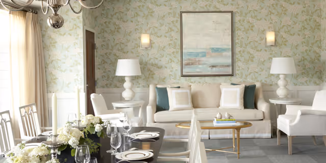 Sunlit, elegantly furnished dining and living area with a set dining table in the foreground, a beige sofa, framed artwork, and decorative lamps.