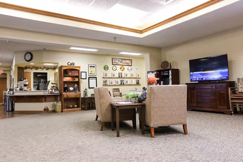 A cozy common area in a senior living facility with two beige armchairs around a small wooden table. In the background, there is a café counter with shelves holding coffee supplies and a wall decorated with plates and framed pictures. A large flat-screen TV is mounted on a wooden cabinet to the right.