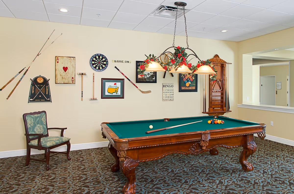 A recreational room with a classic wooden pool table with green felt, a pool cue resting on it, and a set of billiard balls arranged. Above the table hangs a light fixture decorated with red flowers and green leaves. The wall behind the pool table features various game-themed decorations including fishing rods, a dartboard, a croquet mallet, a hockey stick, framed pictures, and a bingo card. A single upholstered chair with wooden arms and legs is positioned to the left. The room has patterned carpet and light yellow walls.