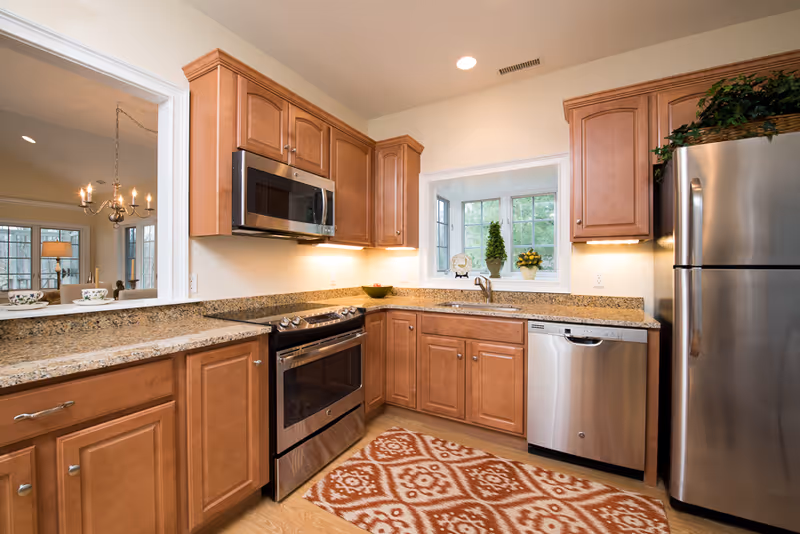 A modern kitchen with wooden cabinets, granite countertops, stainless steel appliances including a refrigerator, dishwasher, microwave, and oven. There is a window above the sink with decorative plants on the windowsill and a patterned rug on the wooden floor. A pass-through window opens to a dining or living area with a chandelier and large windows.