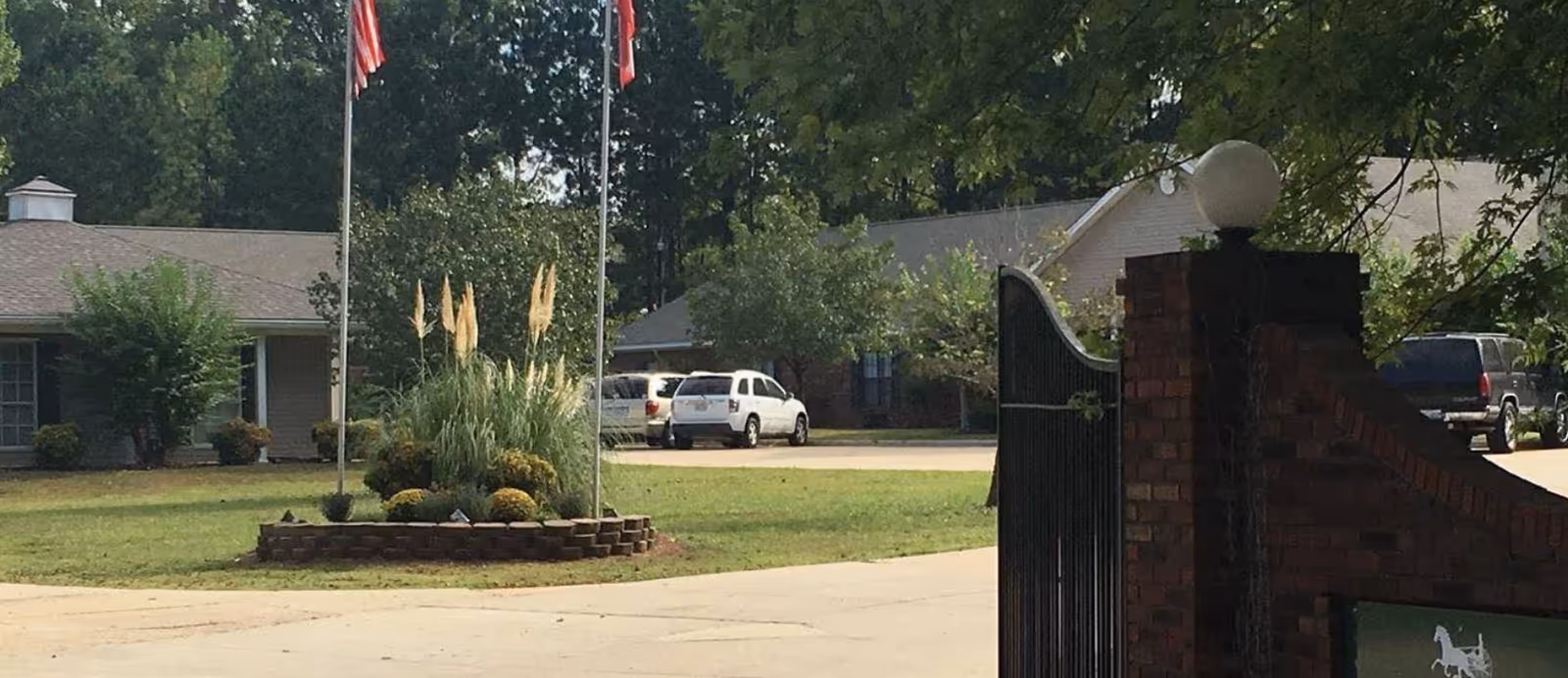 View of the entrance area of Creekside Manor Assisted Living featuring a driveway, landscaped garden with tall grasses and flowers, two flagpoles with flags, and residential buildings in the background surrounded by trees.