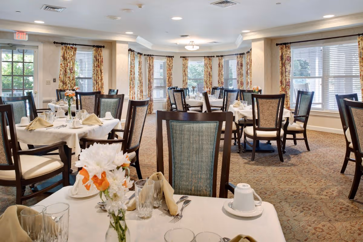 Bright, neatly arranged dining room with round tables set with white tablecloths, napkins, and floral centerpieces.