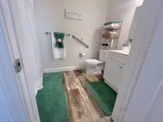 A clean and well-lit bathroom featuring a white toilet, white vanity with a sink, and a metal shelving unit holding towels and toiletries. The floor has wood-like tiles with green bath mats placed near the shower and vanity. A white towel with a green towel is hanging on a wall-mounted towel rack, and a grab bar is installed on the wall next to the toilet.