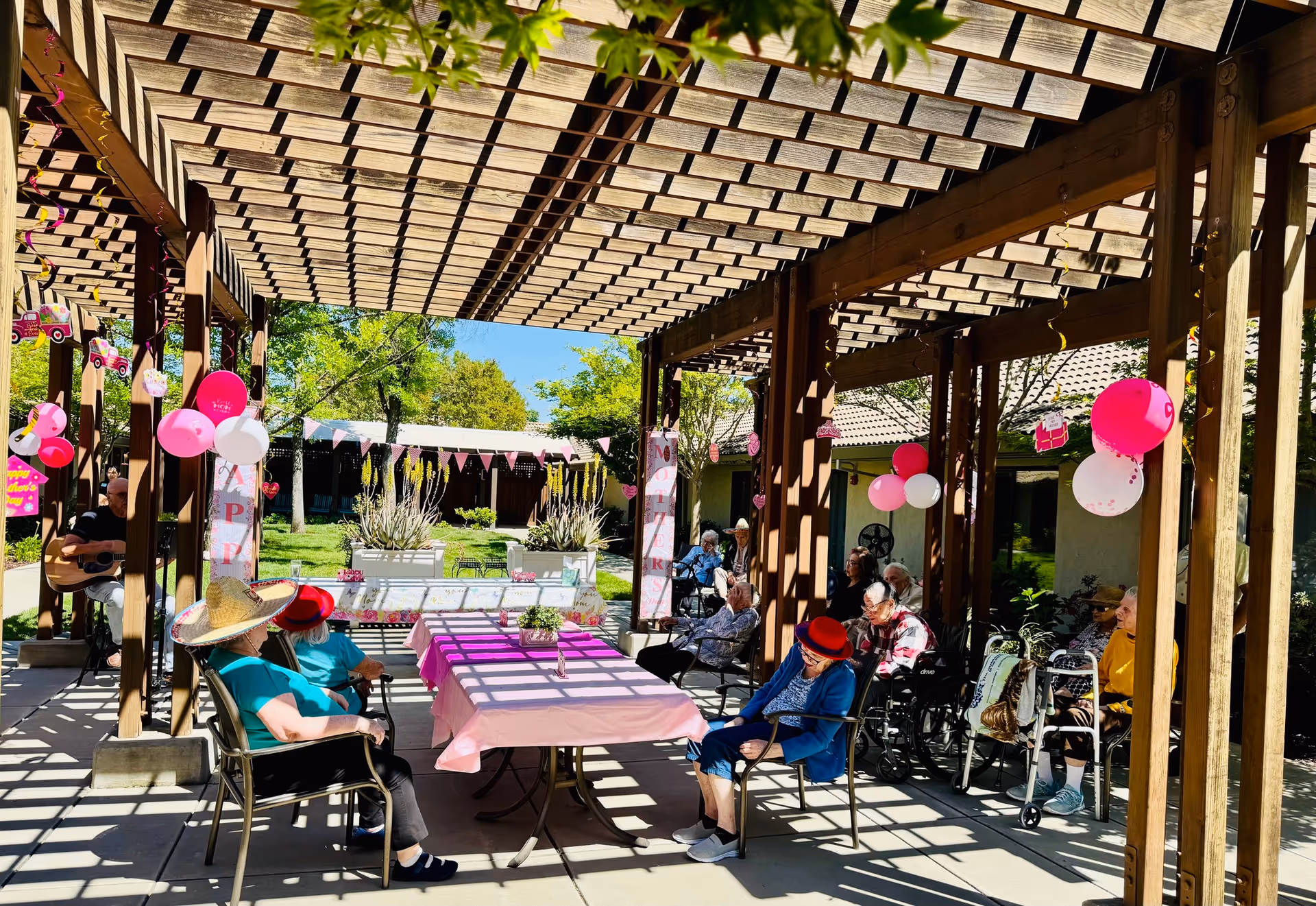 A group of elderly people sitting under a wooden pergola decorated with pink and white balloons and banners. Some are seated in chairs and wheelchairs around a table covered with a pink tablecloth. The setting is outdoors with green trees and a building visible in the background. A person is playing guitar on the left side of the image.