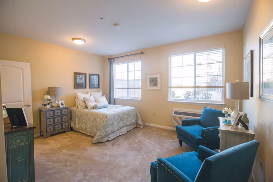 A bright and cozy bedroom with a bed covered in light-colored bedding, two blue armchairs, a small nightstand with a lamp and decorative items, two large windows letting in natural light, and framed artwork on the walls.