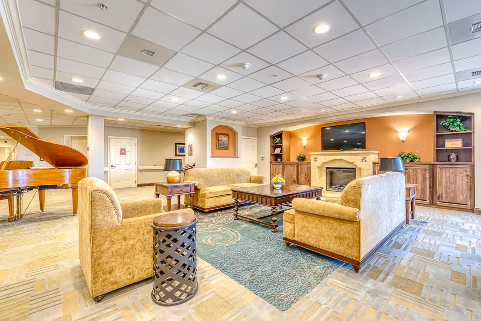 A bright and spacious living room area in a senior living facility featuring beige upholstered sofas and armchairs arranged around a wooden coffee table on a patterned rug. The room includes a fireplace with a mounted flat-screen TV above it, built-in wooden shelves with decorative plants, and a grand piano in the background. The ceiling has recessed lighting and the walls are painted in warm tones.