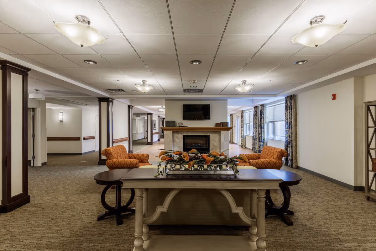 A spacious and well-lit common area with a central fireplace and a mounted TV above it. In front of the fireplace, there is a decorative table with a floral arrangement. Two orange patterned armchairs are positioned on either side of the fireplace. The room has carpeted floors, white walls, and multiple ceiling lights. Large windows with patterned curtains allow natural light to enter the space.