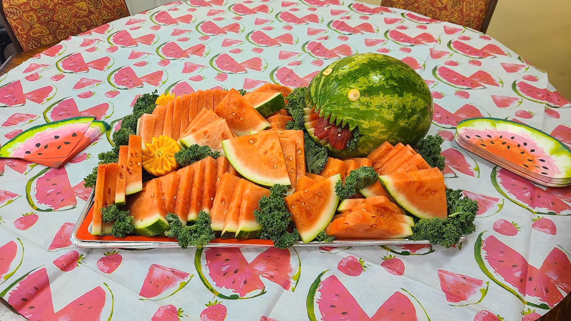 A table covered with a watermelon-themed tablecloth and matching watermelon-shaped napkins. On the table is a tray filled with neatly arranged slices of watermelon and a whole watermelon carved to resemble a fish with strawberry teeth, surrounded by green leafy garnish.