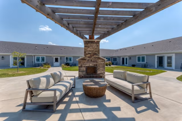 Outdoor courtyard area at Cedarhurst Senior Living of Springfield featuring two cushioned sofas facing each other with a round wicker coffee table in between, under a wooden pergola. A brick fireplace is centered behind the seating area, and single-story building wings with multiple doors and windows surround the courtyard under a clear blue sky.