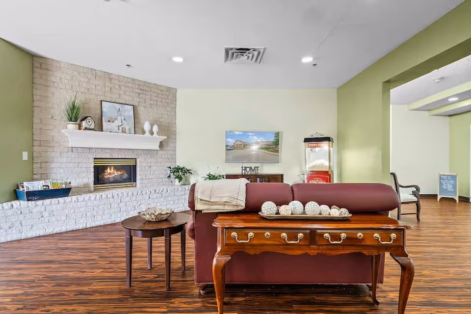 A cozy living room area with a maroon sofa facing away from the camera, a wooden table with decorative balls on it behind the sofa, and a round side table with a decorative bowl. There is a fireplace with a white mantel on a light brick wall to the left, decorated with a plant, clock, and framed picture. A popcorn machine and a TV mounted on a light green wall are visible in the background. The room has wood flooring and recessed ceiling lights.
