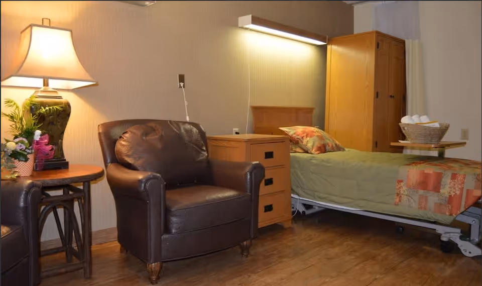A cozy room with a hospital-style bed covered with a green blanket and a colorful pillow. Next to the bed is a wooden nightstand with three drawers and a wall-mounted light above it. There is a brown leather armchair beside a wooden side table with a lamp and a small flower arrangement. A wooden wardrobe is visible in the background.