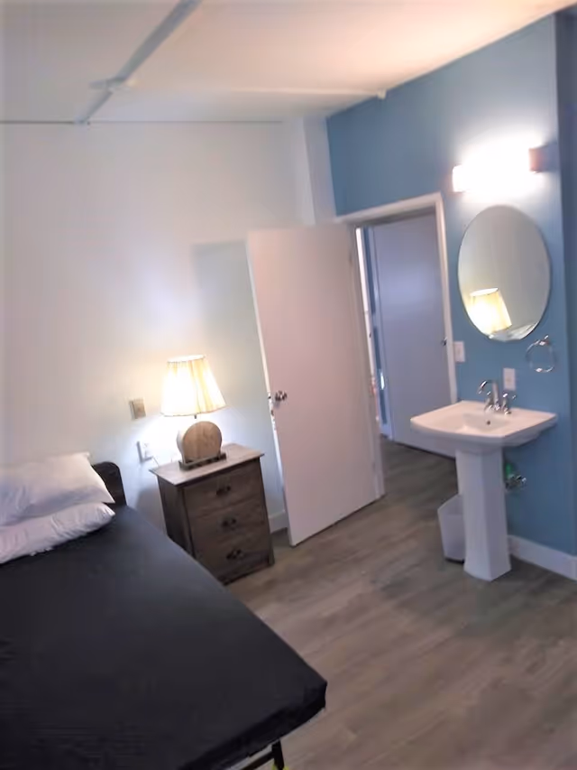 A small bedroom with a single bed covered in a black sheet and white pillows. Next to the bed is a wooden nightstand with three drawers and a lamp on top. The room has light-colored wooden flooring and a blue accent wall with a white pedestal sink and a round mirror above it. An open door leads to another room with similar flooring.