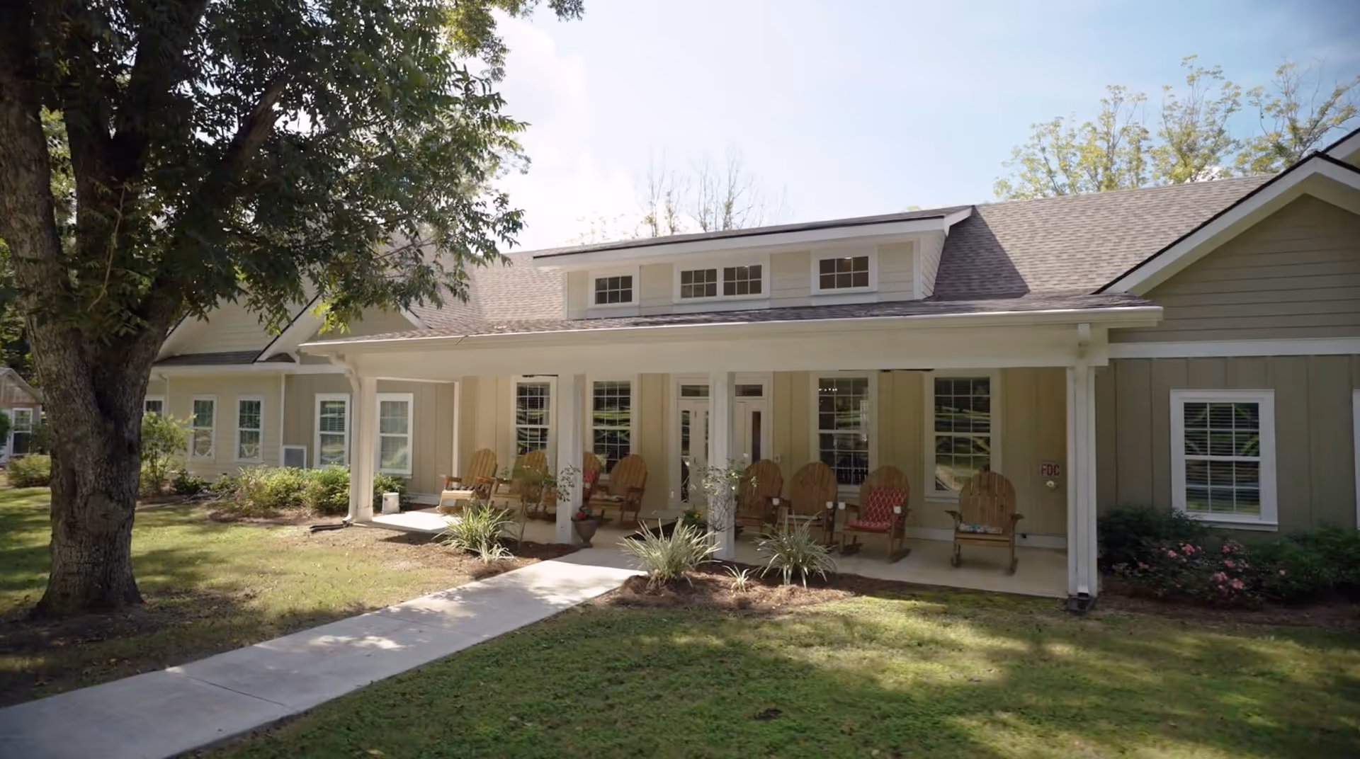 Exterior view of a single-story building with a covered porch featuring several wooden rocking chairs. The building is surrounded by a well-maintained lawn, shrubs, and a large tree providing shade. The sky is clear and sunny.