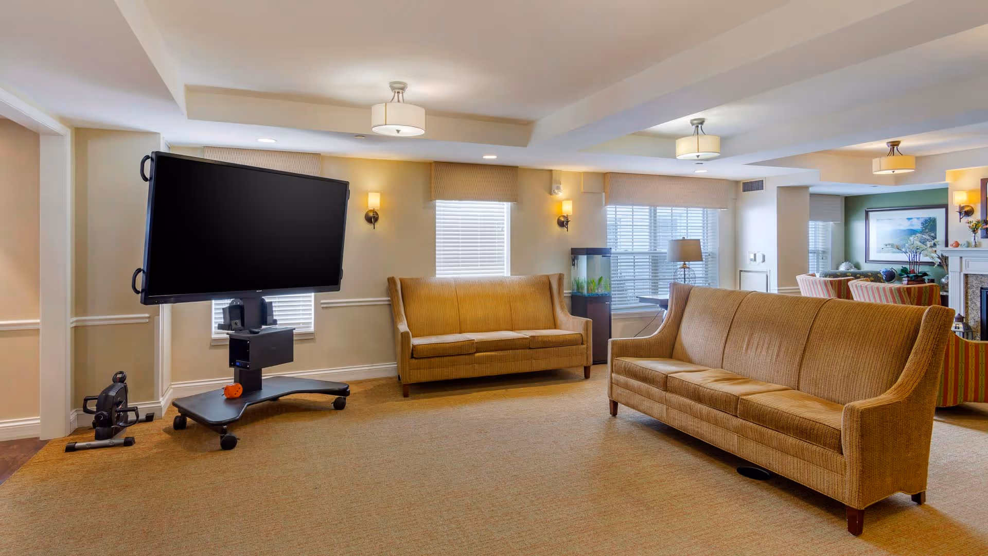 A senior living common room with two tan sofas, a large mobile TV on a rolling stand, windows, and soft overhead lighting.