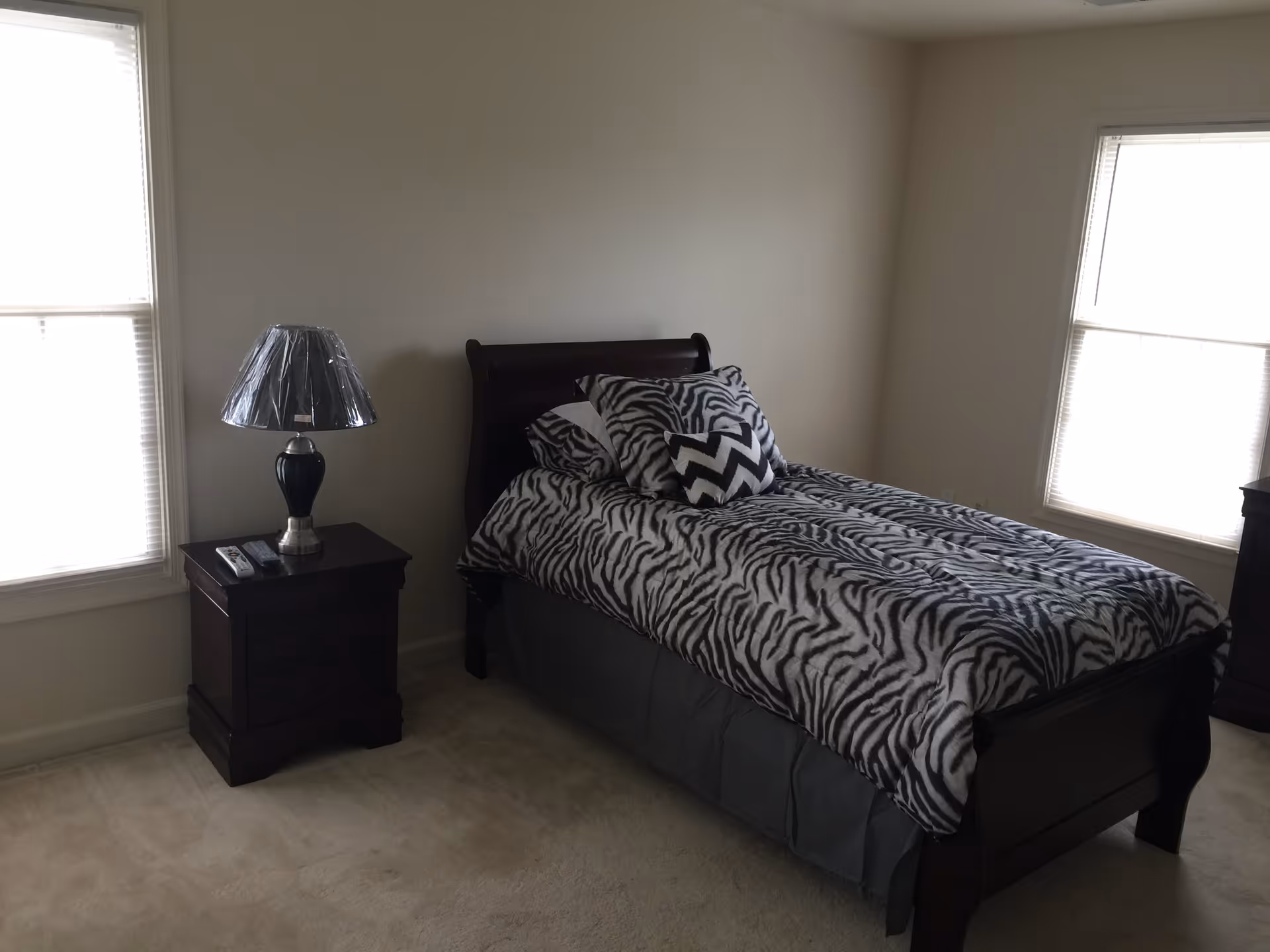 A bedroom with a single dark wooden bed featuring zebra-patterned bedding and pillows. There are two windows with blinds on either side of the bed. A dark wooden nightstand with a lamp covered in plastic is positioned next to the bed.