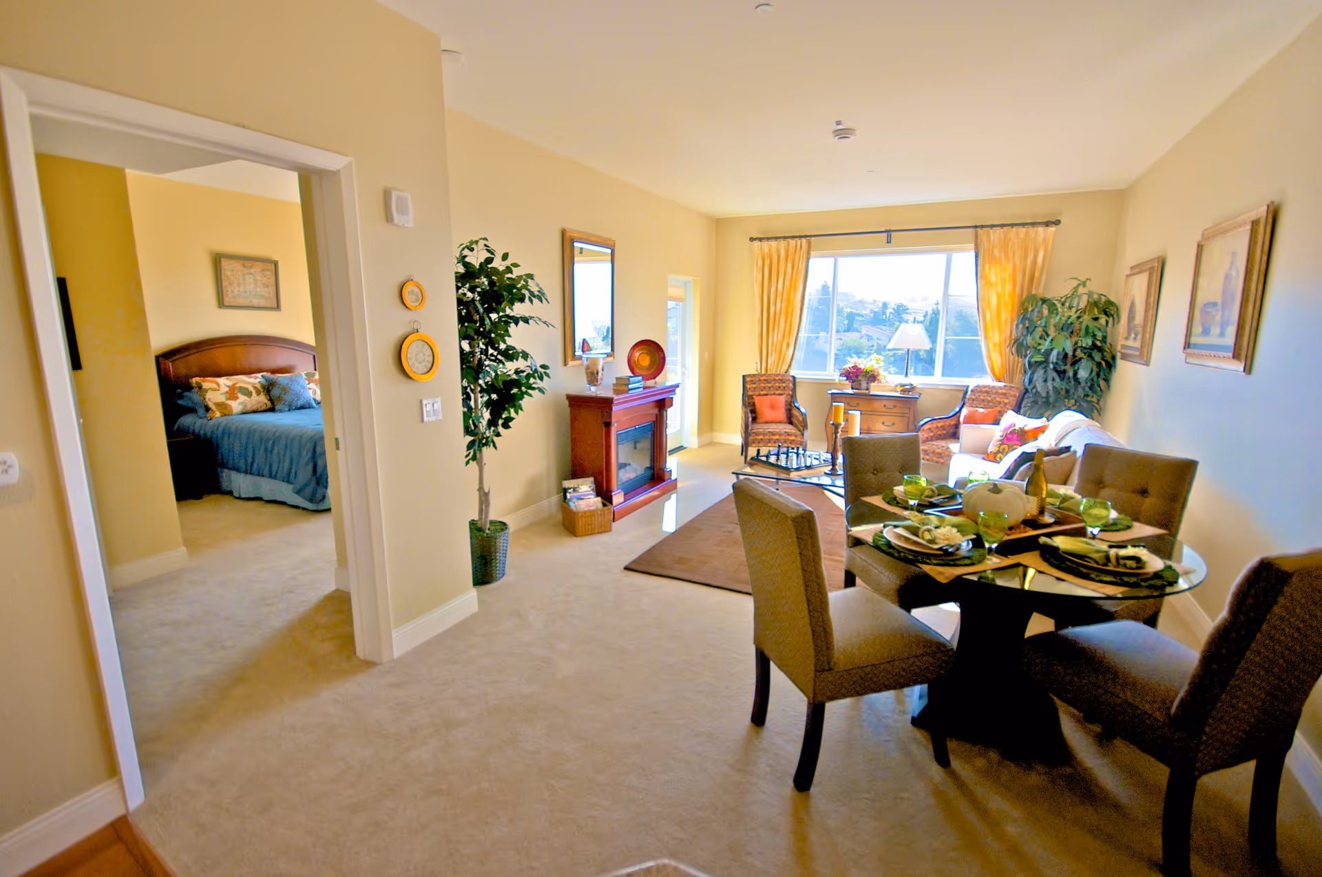 A bright and cozy living and dining area in a retirement community apartment. The room features a round glass dining table set for four with green glasses and plates. The living area has a sofa, two armchairs, a coffee table with a chess set, a wooden fireplace, and large windows with yellow curtains letting in natural light. A doorway leads to a bedroom with a bed covered in blue bedding and decorative pillows. The walls are painted beige, and there are framed pictures and plants adding to the decor.