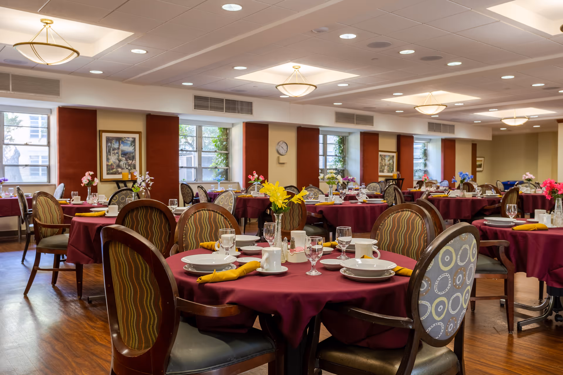 A spacious dining room with multiple round tables covered in burgundy tablecloths, each set with plates, cups, glasses, and yellow napkins. The chairs have patterned upholstery, and there are flower arrangements on each table. The room has large windows letting in natural light, framed artwork on the walls, and ceiling lights providing additional illumination.