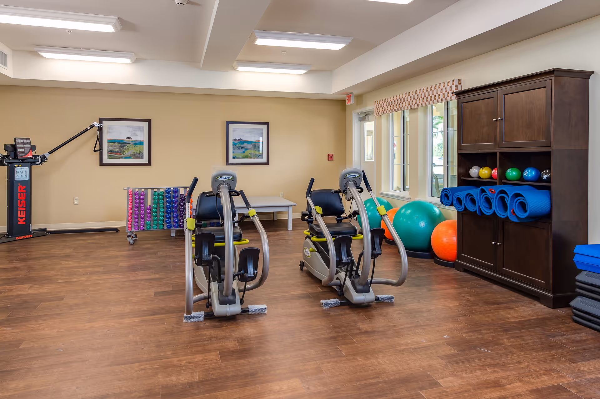 A fitness room with exercise equipment including two recumbent bikes, a Keiser machine, a rack of colorful dumbbells, large exercise balls, and rolled-up yoga mats stored in a wooden cabinet. The room has wooden flooring, beige walls, two framed pictures, and windows letting in natural light.