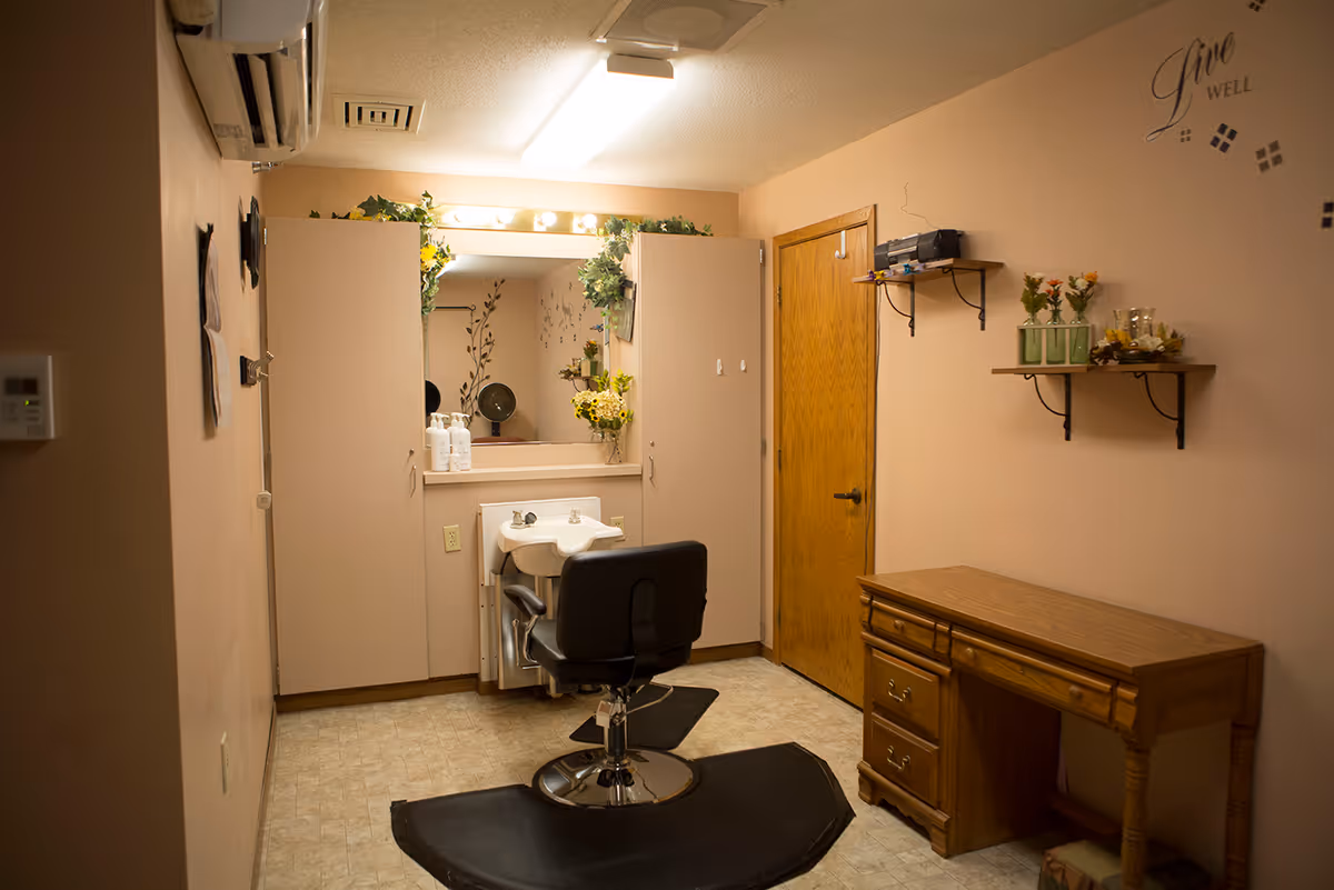 A small room with peach-colored walls featuring a salon chair in front of a sink and mirror surrounded by cabinets. There is a wooden desk with drawers on the right side and two wall shelves with decorative items and flowers. The wall has a decal that says 'Live Well'.