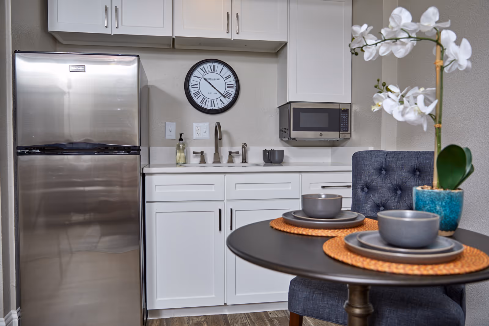 A modern kitchen area with white cabinets, a stainless steel refrigerator, a wall-mounted microwave, and a sink with a faucet. A round table with two place settings and a potted white orchid is in the foreground. A round wall clock with Roman numerals is mounted on the wall above the sink.