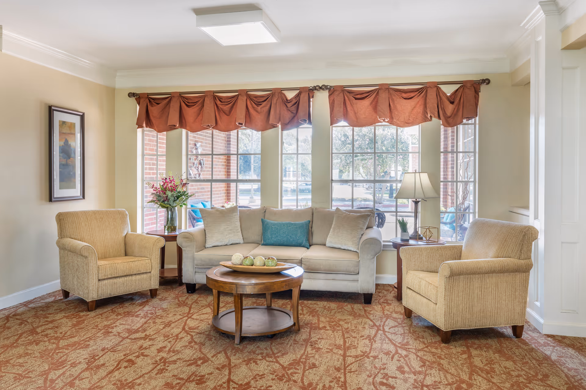 A cozy living room with a beige sofa and two matching armchairs arranged around a round wooden coffee table. The room has large windows with red valance curtains, allowing natural light to fill the space. There are side tables with a vase of flowers and a lamp, and a framed picture on the wall.