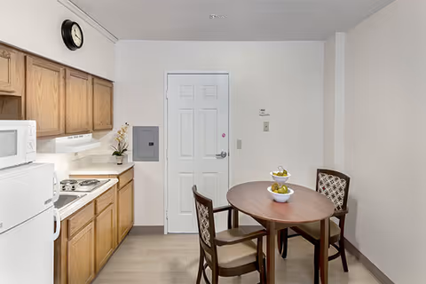 A small kitchen and dining area featuring wooden cabinets, a white microwave, a white refrigerator, a stove with two burners, and a round wooden dining table with two chairs. The table has a two-tiered fruit stand with bananas and apples. A clock is mounted on the wall above the cabinets, and a white door is visible in the background.
