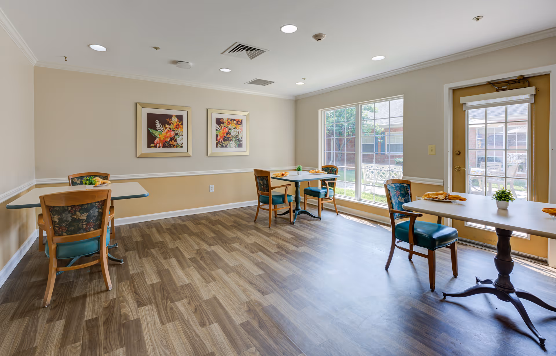 A bright dining room with three tables, each surrounded by wooden chairs with floral upholstery and teal cushions. The room has wood-patterned flooring, beige walls with white trim, two framed floral paintings on one wall, and large windows letting in natural light. A door with glass panes leads outside to a garden area.