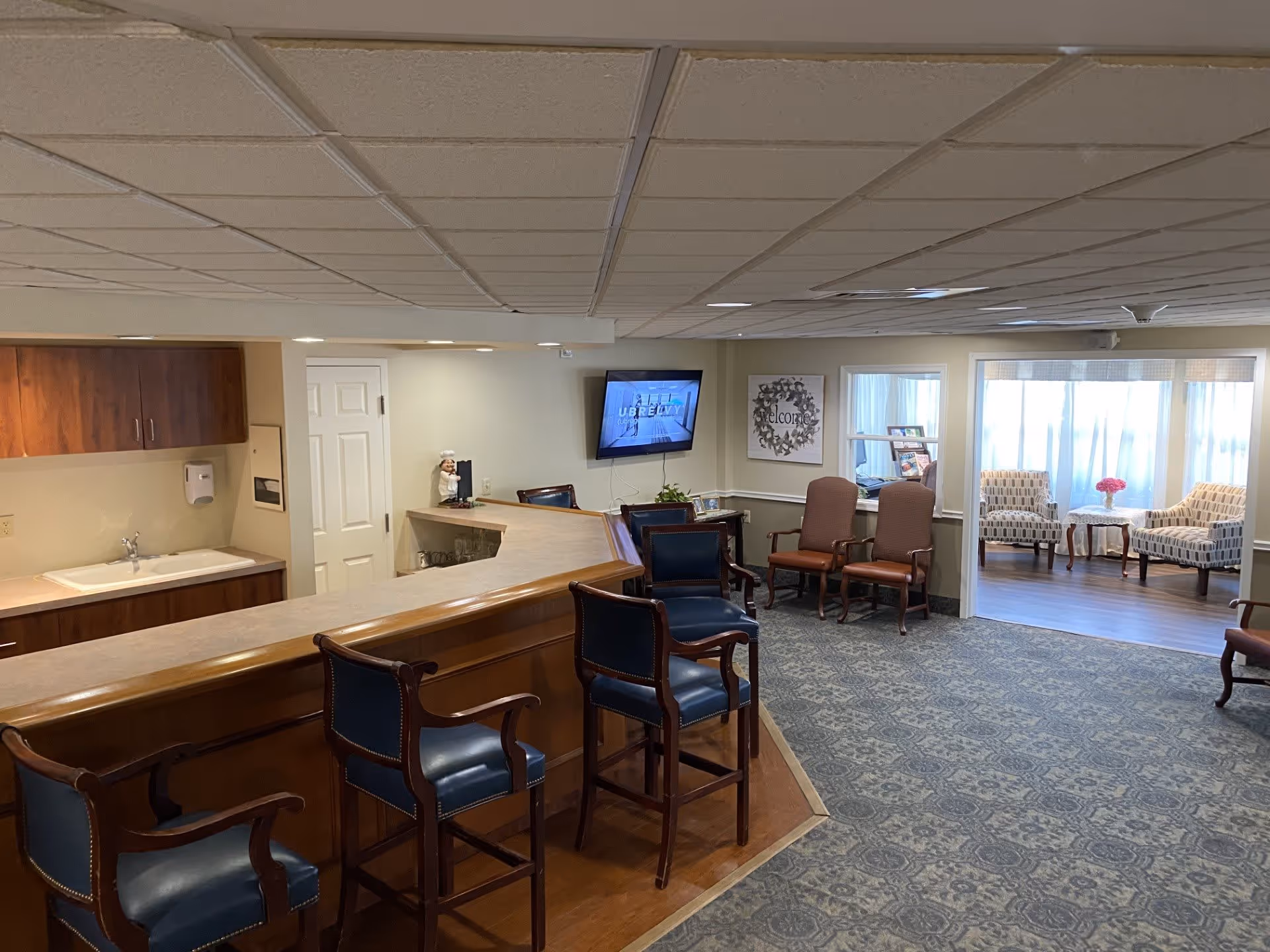Interior of a senior living facility lounge area with a wooden counter and four high chairs with blue cushions. Behind the counter is a sink and cabinets. In the background, there are several chairs arranged around the room, a wall-mounted TV, and a small sitting area with two patterned armchairs and a table with a flower vase near a window with sheer curtains.