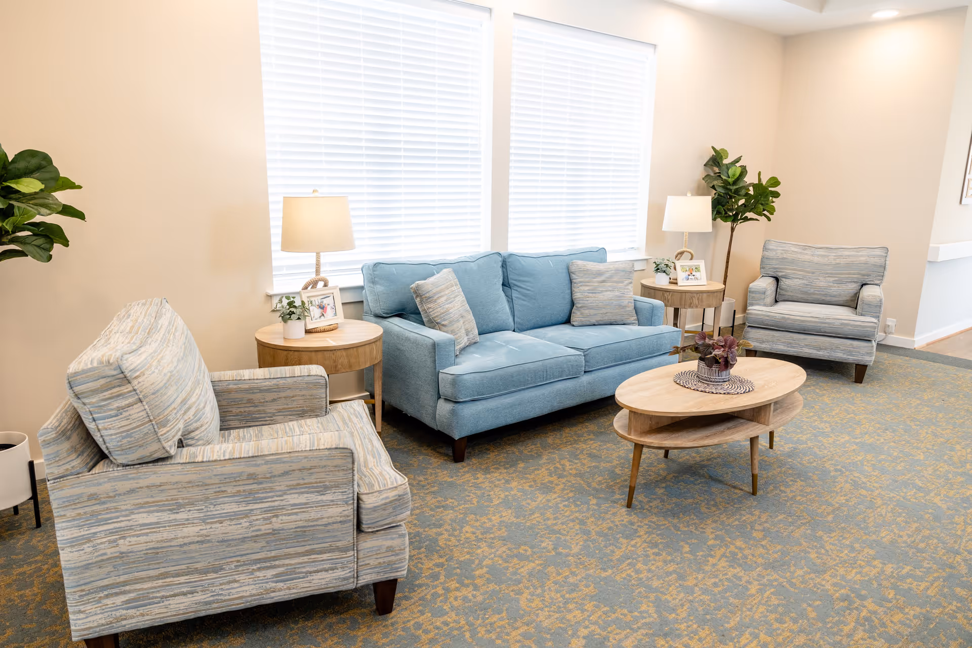 A bright and cozy living room area featuring a blue sofa with two matching pillows, two patterned armchairs, a wooden oval coffee table with a decorative centerpiece, two round wooden side tables each with a lamp and framed photo, and two green potted plants near the windows with white blinds.