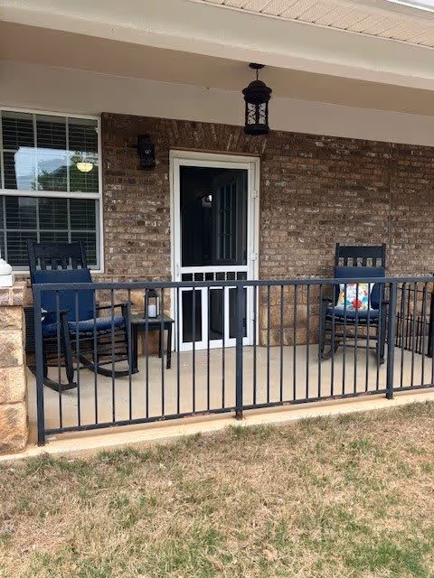 A covered porch area with two black rocking chairs, one with a colorful cushion, and a small side table between them. The porch has a black metal railing and a brick wall with a window and a door leading inside. A hanging lantern light fixture is mounted on the ceiling above the door.