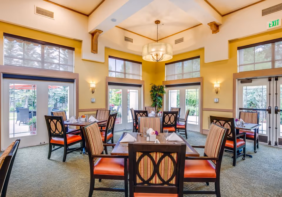 Bright dining room with multiple set tables and upholstered chairs, large windows and glass doors opening to an outdoor patio.