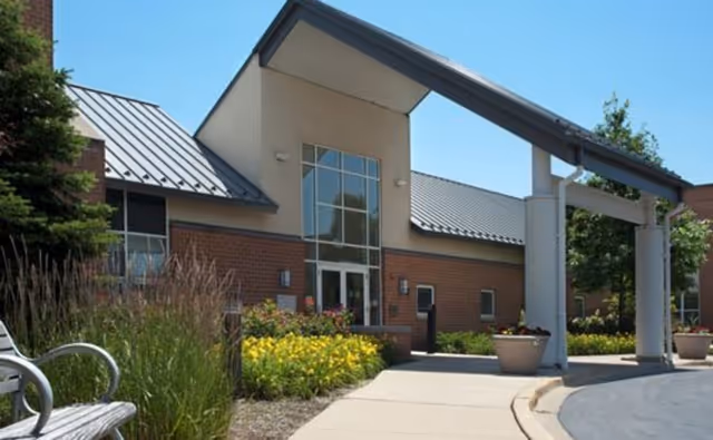 Entrance of a senior living facility with a covered drop-off area, brick and beige exterior walls, large windows, and landscaped garden beds with yellow flowers and greenery under a clear blue sky.