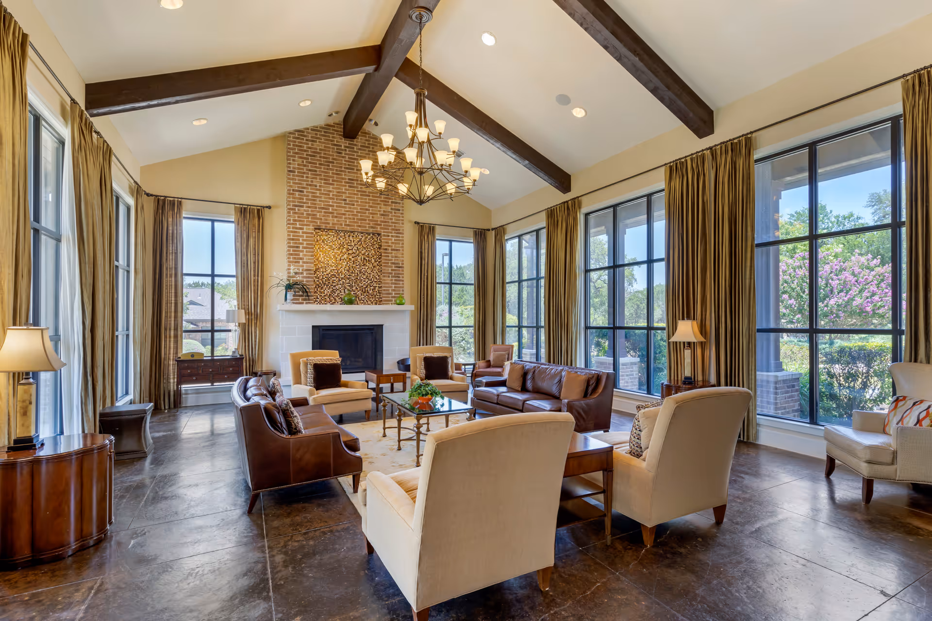 Spacious living room with high vaulted ceiling featuring exposed wooden beams and a large chandelier. The room has a brick fireplace with a decorative panel above it, surrounded by large windows with long beige curtains letting in natural light. The seating area includes a mix of beige and brown upholstered armchairs and sofas arranged around a glass coffee table on a light-colored rug. There are side tables with lamps and a view of greenery and flowers outside the windows.