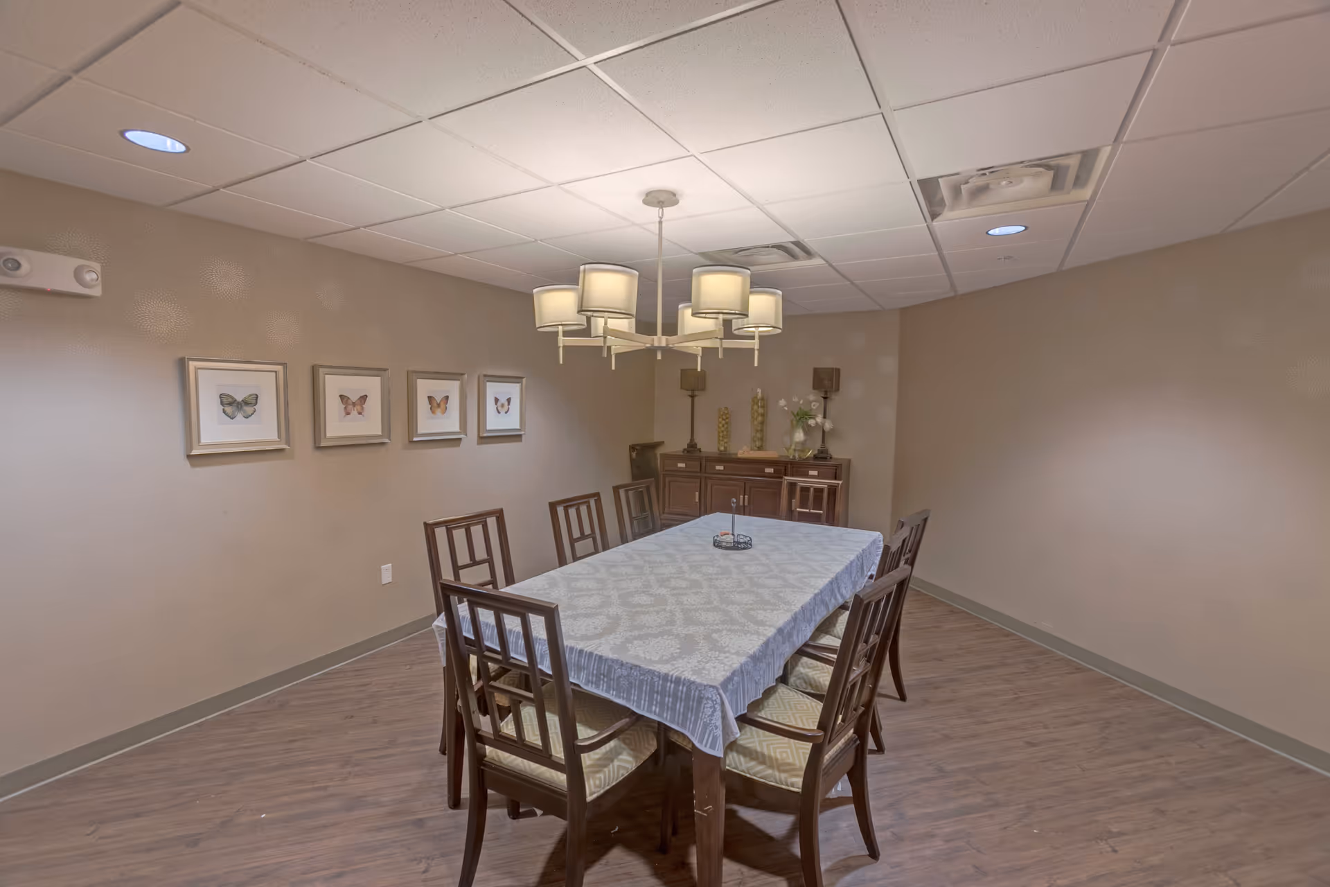 A small dining room with a rectangular table covered by a tablecloth, eight wooden chairs, an overhead chandelier, and a sideboard against beige walls with framed butterfly prints.