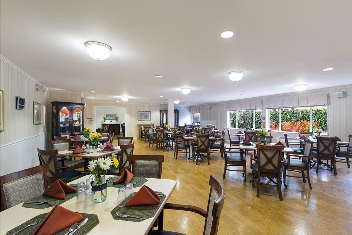 A spacious dining room with multiple tables set with green placemats, folded rust-colored napkins, silverware, and glasses. Each table has a vase with fresh flowers. The room has wooden floors, light-colored walls with framed artwork, and large windows letting in natural light. There are several chairs around each table and a cabinet with glass doors in the background.