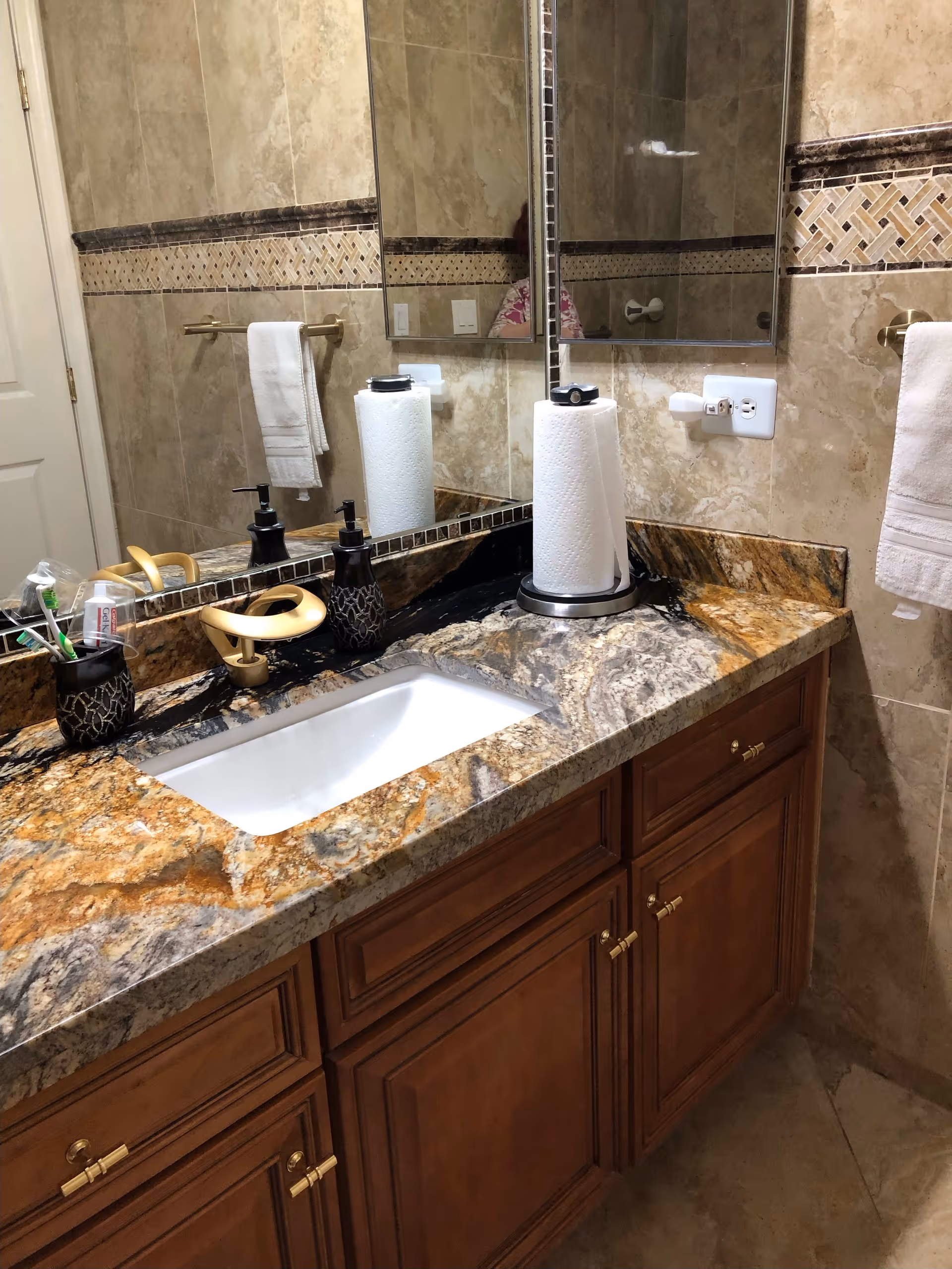 A bathroom countertop with a built-in sink, a gold-colored faucet, a soap dispenser, a toothbrush holder with toothbrushes, and a paper towel roll. The countertop is made of a brown and beige marble pattern. The wall behind has beige tiles with a decorative mosaic tile border. A large mirror is mounted above the sink, reflecting part of the bathroom. A white towel hangs on a gold towel rack on the right side.
