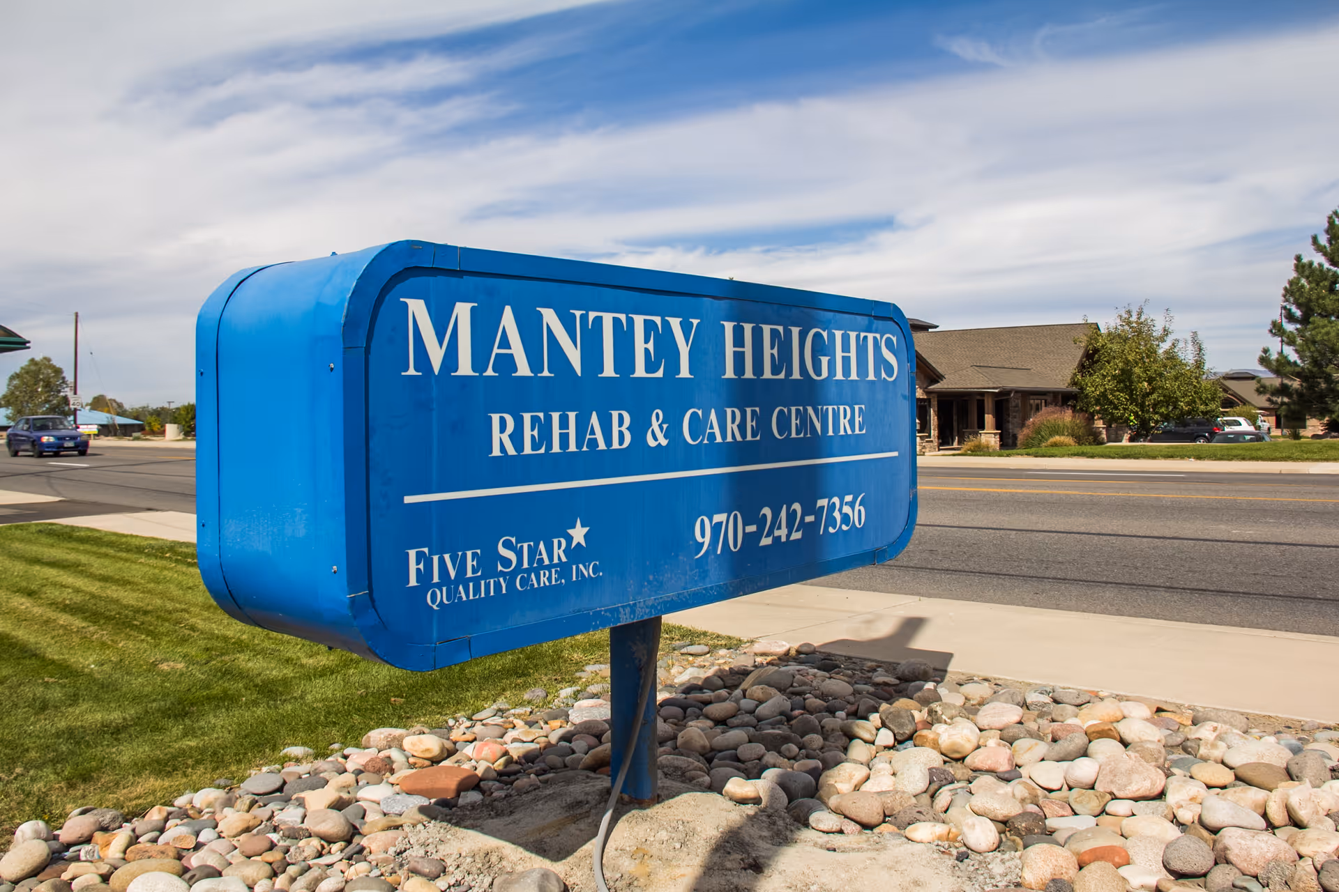 Blue sign for Mantey Heights Rehab & Care Centre with contact number 970-242-7356, located on a roadside with grass and rocks around the base of the sign and a building in the background.