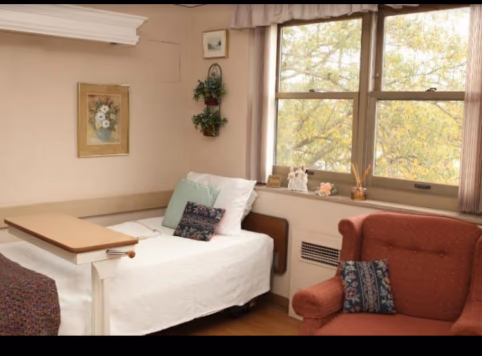 A cozy bedroom with a single bed covered in white bedding and decorative pillows. Next to the bed is a wooden overbed table. A red upholstered armchair with a patterned pillow sits nearby. The room has a large window with a view of trees outside, and the walls are decorated with framed pictures and hanging plants.