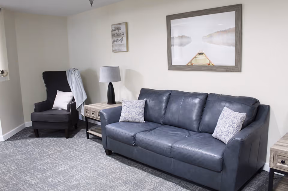 A furnished living room with a gray leather sofa, patterned pillows, an upholstered armchair, side tables, a lamp, and framed wall art.
